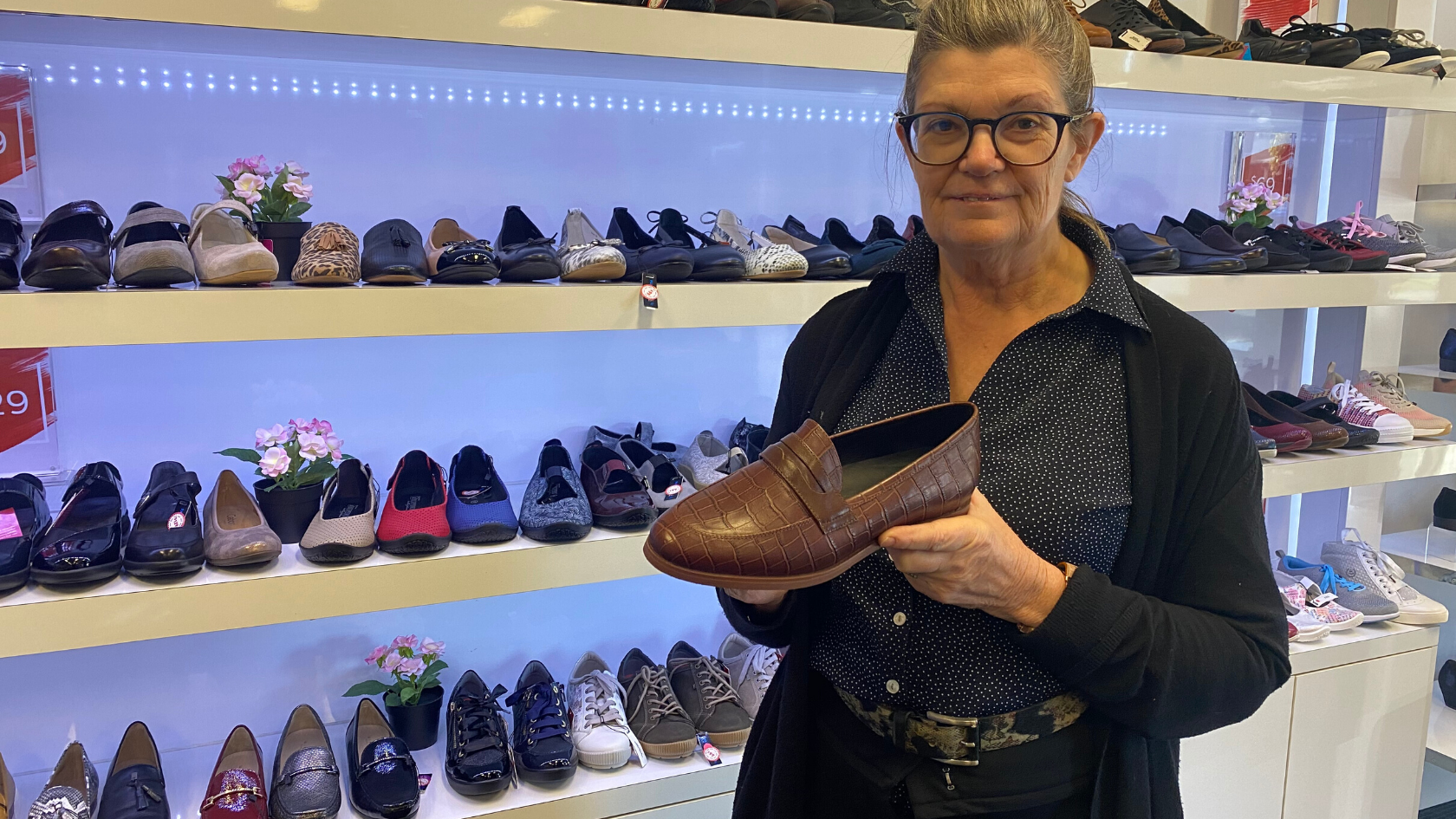 An older woman holding a brown leather loafer in front of a shoe display.